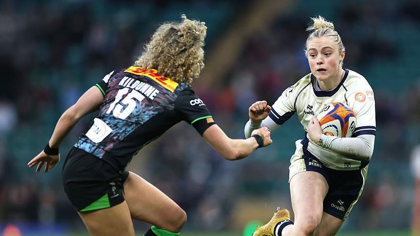 Millie David (right) runs with the ball in her left arm with her right held out in a fist as Ellie Kildunne of Harlequins (left) runs towards her during a match