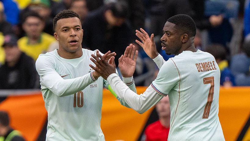 Kylian Mbappe (left) and Ousmane Dembele (right, photographed from behind), both wearing white France shirts, high five one another