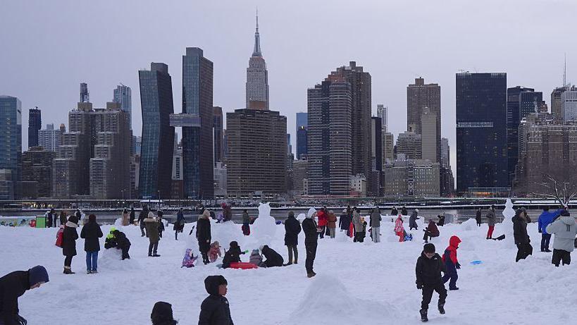 Families with young children are playing in the snow. Skyscrapers are behind them.