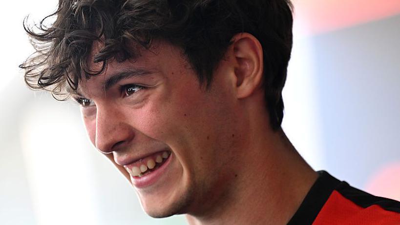 Haas racing driver Ollie Bearman smiling at the camera. He's facing to the side so you can see the profile of his face. He has brown eyes and brown curly hair. He's wearing a red t-shirt with a black collar