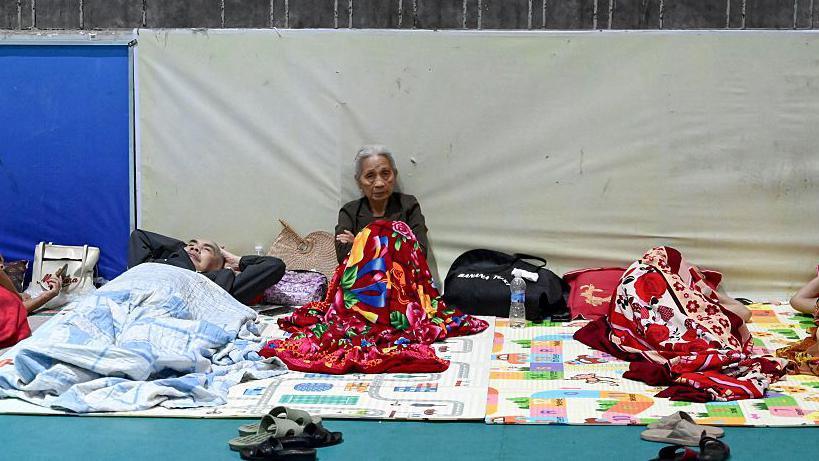 An old lady sits on the floor of an evacuation centre, covered by blankets and mats around her