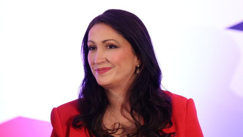 Pengelly has long black hair and is wearing a red blazer. She is wearing makeup and has earrings in. Behind is lit-up blurred background.