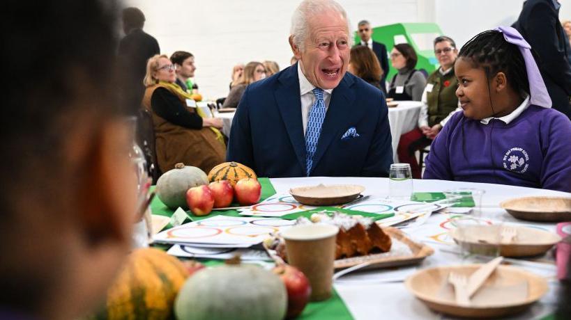 A laughing Prince Charles seated at a table with a smiling schoolgirl wearing a purple school uniform sweatshirt to his left. On the table are plates with knives and forks and a collection of apples and various squashes. In the foreground is the blurred profile of another child.