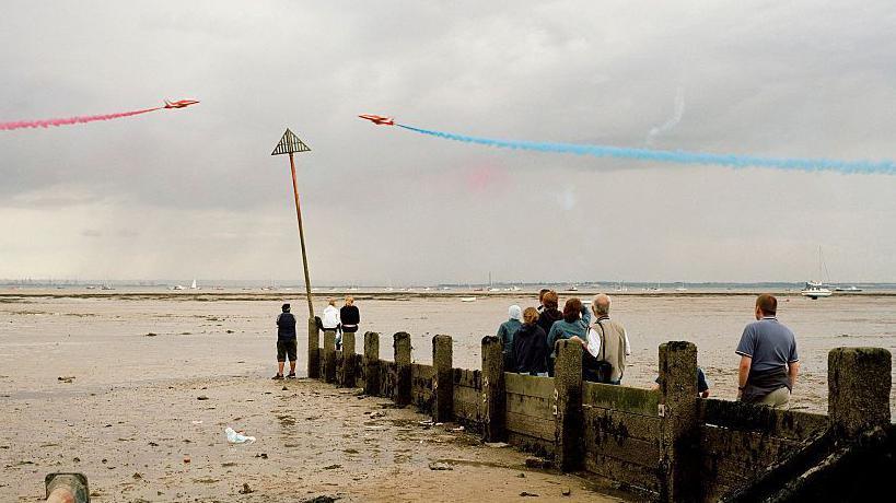 The Red Arrows flying above the water, which is at low tide, in Southend-on-Sea. One of the jets is blasting out a trail of red smoke, while the other has blue smoke. A small crowd of people are standing on mud watching them fly past.