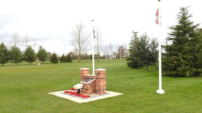 A memorial bearing the details of the airmen who died, with a British and Canadian flag flying at either side, on a green lawn on a golf course. There are poppy wreaths on the ground in front of the memorial.