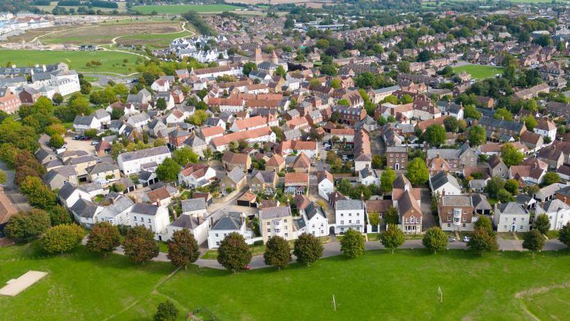 An aerial view of part of Poundbury in 2022. Well-established trees in close proximity to each other line a road, which divides houses from an area of grass.