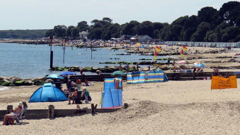 A curved sandy beach on a bright, breezy day. There are rock groynes, lifeguard flags and several beachgoers with portable shelters and chairs.