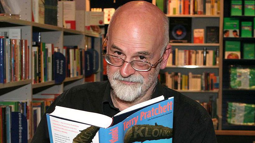 Sir Terry reading the hardback version of his book Klonk which is in a blue dust cover. He is sat in a library and surrounded by books.