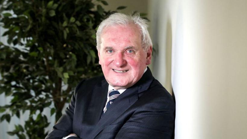 A man with short grey hair and red skin smiling at the camera as he poses. He is wearing a white shirt, black suit jacket and striped dark tie.