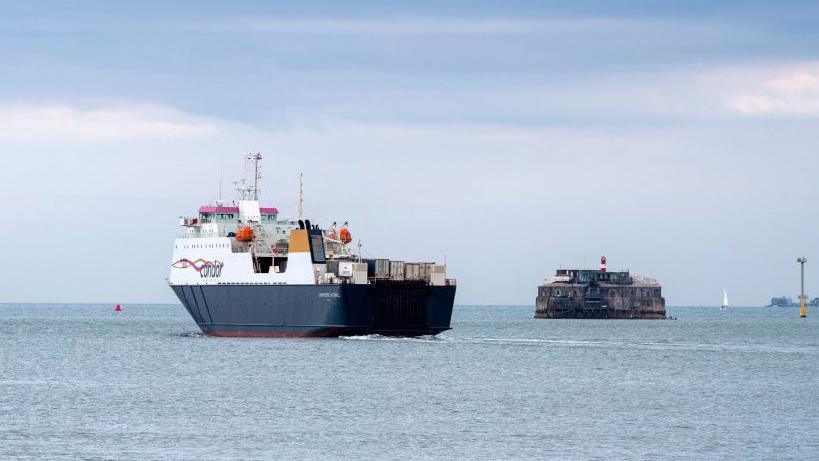 A photo of Spitbank Fort from a little further away, with a large ferry going past it. Other boats and some buoys can be seen around it.