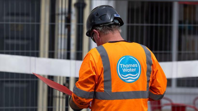 A Thames Water employee wearing a hi-vis uniform reads a document inside a red folder while on a site