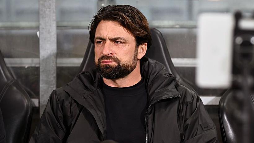 A bearded Russell Martin sitting on a coaching bench wearing a black round-neck sweater and black zipper jacket