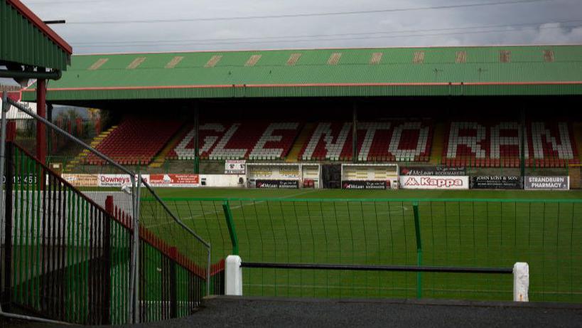 Glentoran's Oval stadium sits empty on a cloudy day. The green roofed stand has red seats with 'Glentoran' spelt out in white seats. The football pitch is surrounded by a green fence.