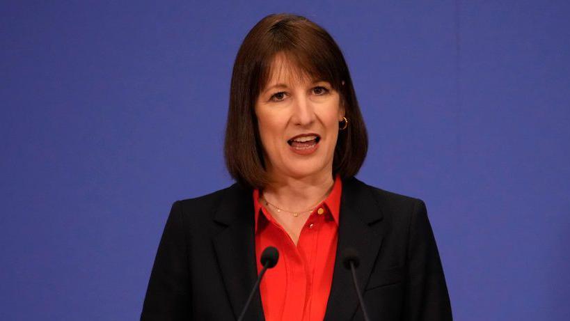 Rachel Reeves wearing a red shirt and black blazer. She is talking with two small mics in front of her. The background is blue.