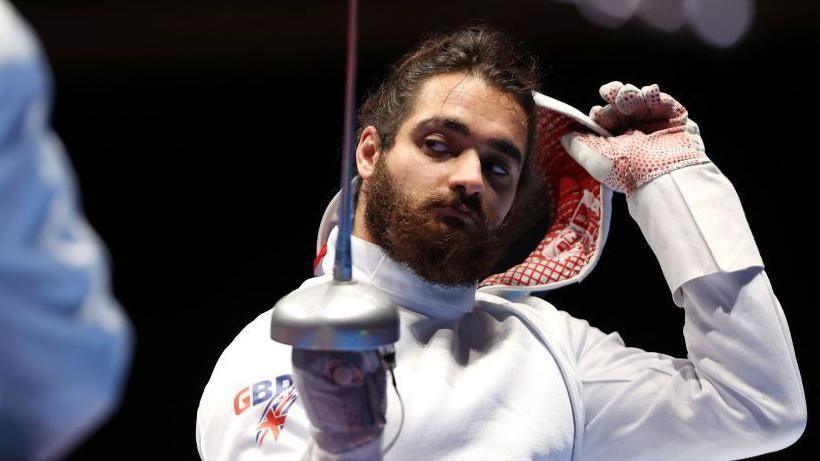 Dimitri Coutya taking off his fencing hood and looking at his opponent while holding up his fencing foil (sword). He is dressed in all white protective kit with gloves. There is the Great Britain logo on his shoulder.