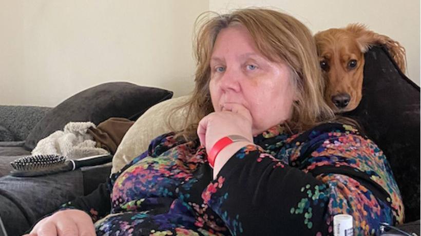 A woman sat on a sofa with her finger in her mouth and a dog resting behind her head.