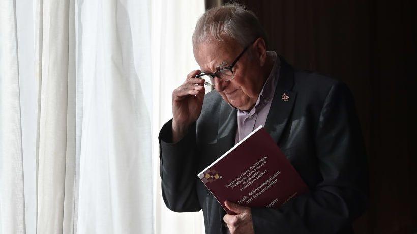 Professor Phil Scraton is seen clutching a report, helium  has achromatic  hairsbreadth  and is holding his glasses.