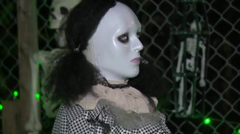 A mannequin wearing a black and white frilly dress, black wig and dark makeup stands in a front garden. 