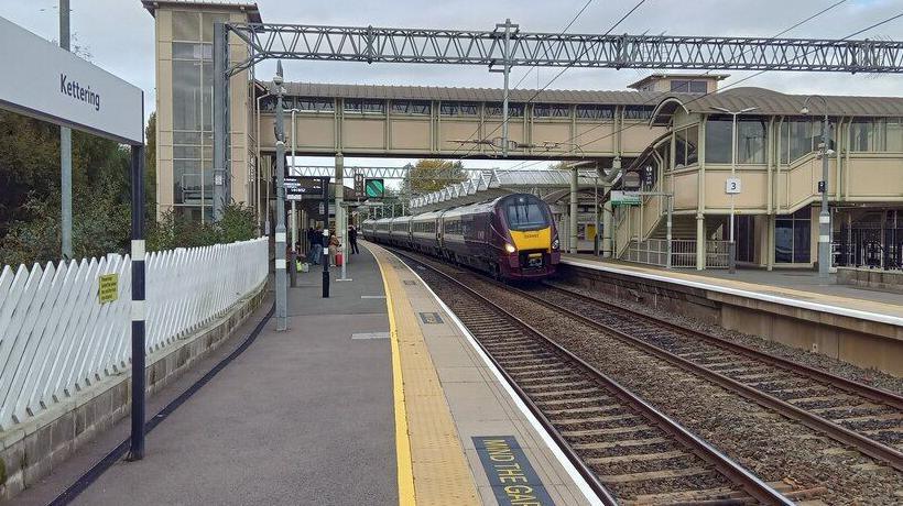 Railway platform with train approaching. A few people can be seen in the distance of the platform and a sign saying Kettering is at the front of the image.
