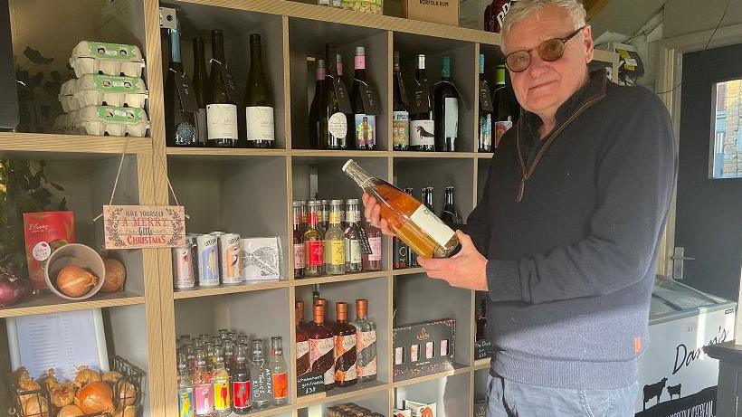 Neil Shearer holds a bottle of wine at Stanhoe Stores. He is wearing a blue half zip jumper and blue chinos. He has white hair and is wearing glasses. 