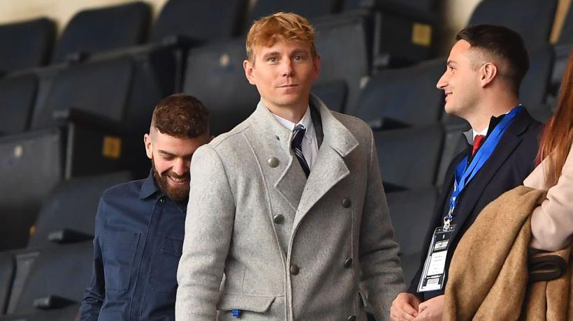 Dane Murphy dressed in a white shirt with grey, striped tie and a light grey overcoat in a football stand