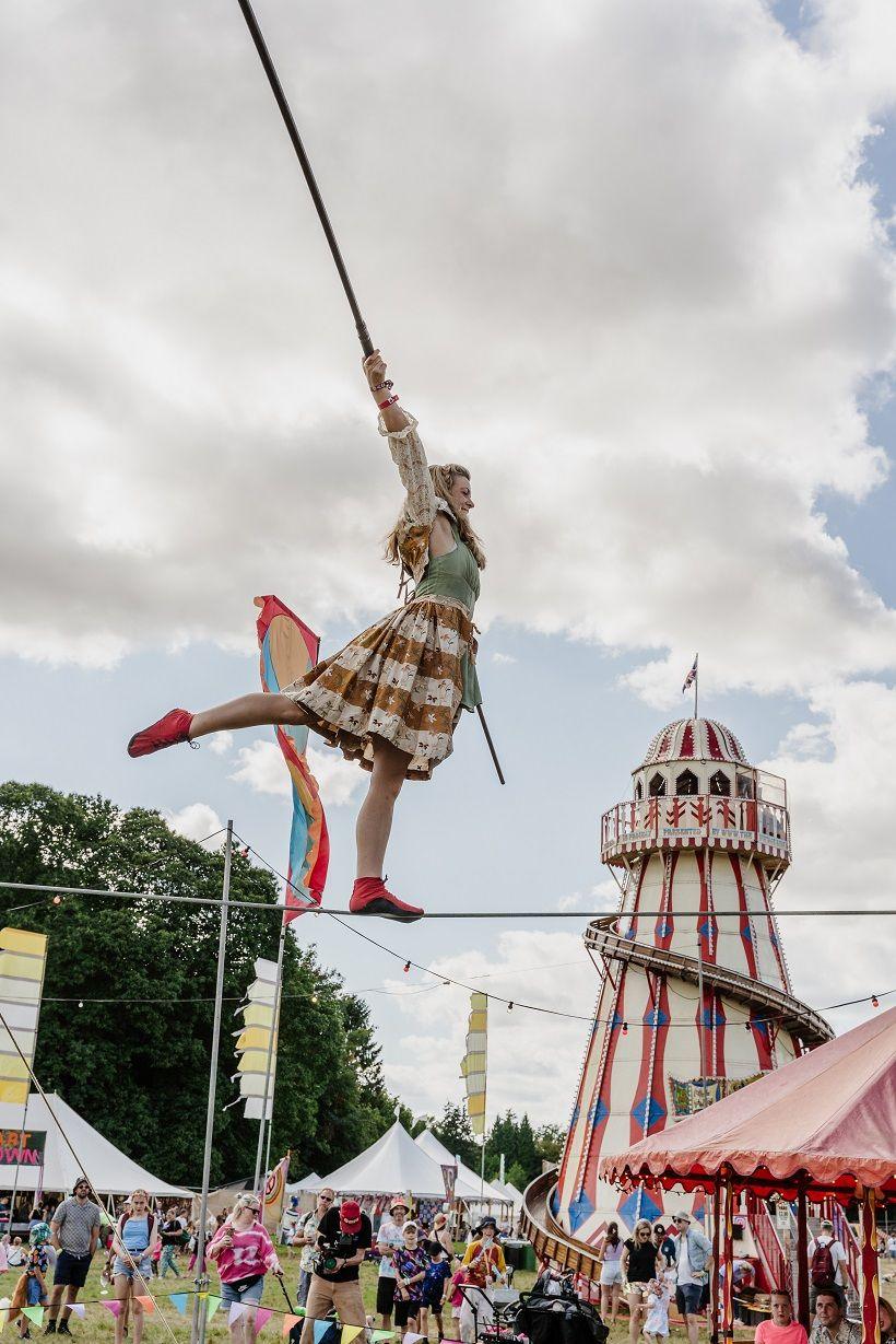 A woman walks on a tightrope while holding a pole for balance. A crowd is beneath her in front of a helter-skelter ride. 