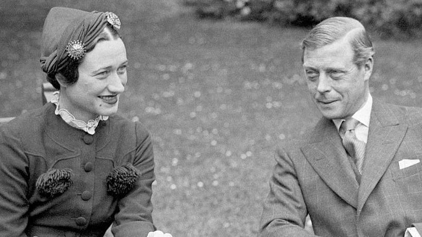 Wallis Simpson sits with Edward VIII in a garden. They are smiling and look away from the camera. It is a black and white image.