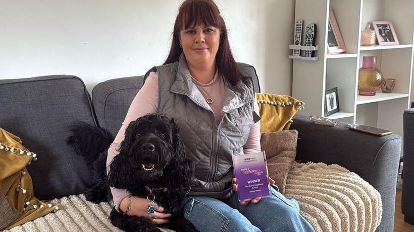 Ashley Timms, who has red/brown hair, sits with her black dog on her sofa. She is wearing jeans, a pink top and a grey gilet. She is holding her purple Make A Difference glass award with certificate.