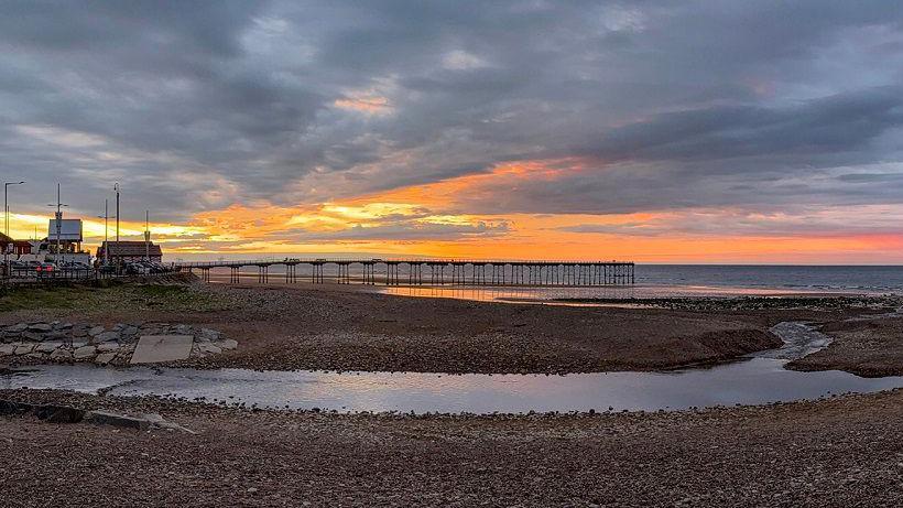 Saltburn beach which is covered in pebbles. A large pool of water is flowing into the sea through the middle of the beach. A pier can be seen in the distance. The sky is grey and orange.