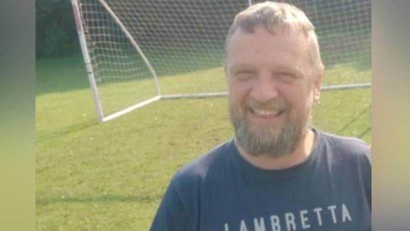 Robin Reed smiling for the camera. He has a grey beard and is wearing a navy blue t-shirt that has the Labretta brand. Behind him is a football goalpost.