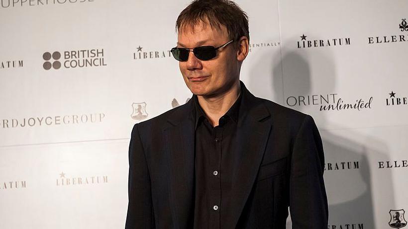 Orbit wearing a black suit and sunglasses, slightly smiling on a red carpet