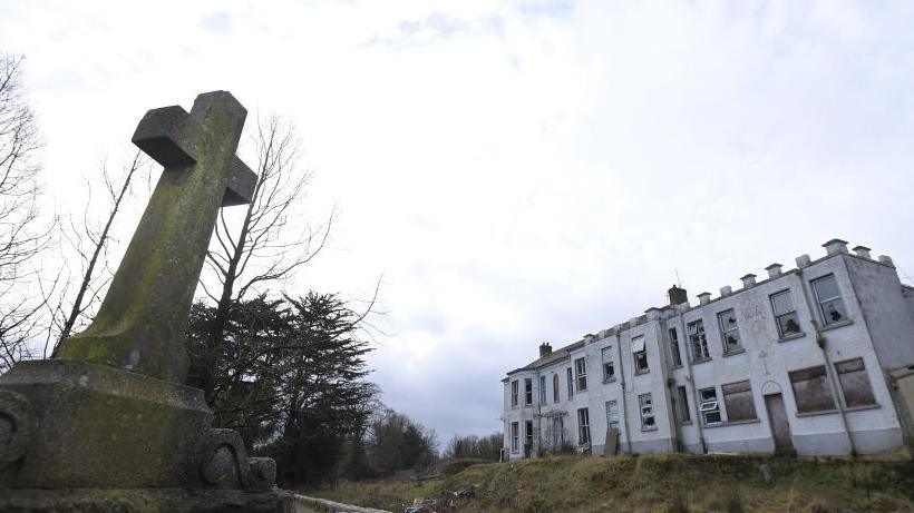 The former Marianvale Mother and Baby home in Newry. A dilapidated two-storey building surrounded by overgrown grounds and a holy cross plinth.