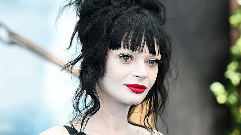 Ruth Codd has pale, white make-up covering her neck and shoulders. She has bright red lipstick and dark mascara. She is smiling slightly, looking vaguely towards the camera, with a light blue and black backdrop at a press event for a recent film release.