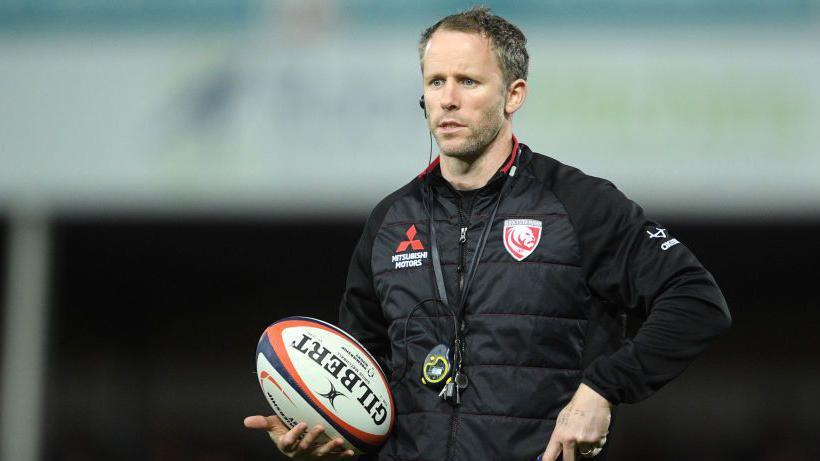 Dan Tobin holding a rugby ball while wearing a Gloucester jacket