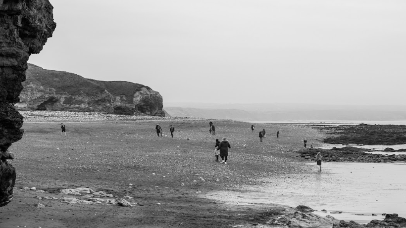 A black and white photo of people walking around Blackhall Colliery beach in the distance looking at the sand. A cliff can be seen in the background.