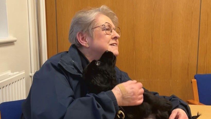 Woman in blue jacket cuddles a black puppy on a table. She has short grey hair and is wearing glasses. The puppy has its head angled towards her face