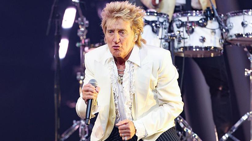 Sir Rod Stewart pouts on stage while holding a microphone.