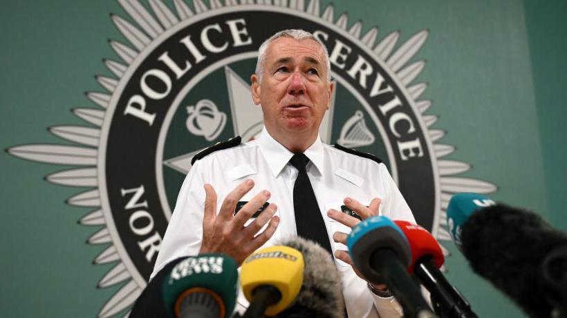 Shows Jon Boutcher in a white shirt and black tie standing in front of the Police Service of Northern Ireland crest. 