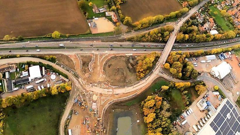 A birds-eye-view image of the roadworks. It shows a new junction that is being built above two busymain roads. To the left there is a large building with solar panels.