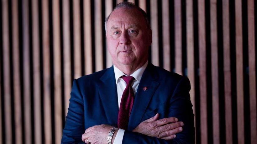 Managing director of Mineral Resources Chris Ellison wearing blue suit, white shirt, and red tie with silver watch folding arms in front of wooden background.