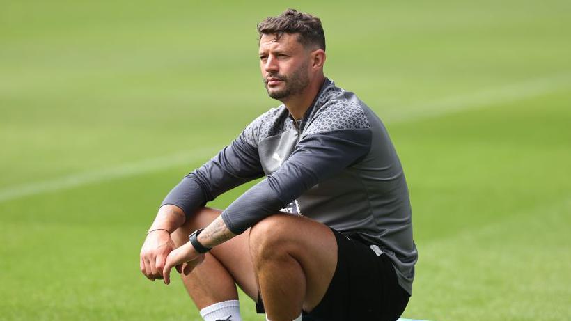 A man in grey and black football training kit sitting on a pitch.