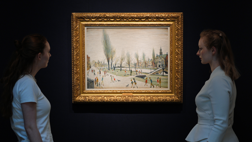 Two women in white t-shirts stand either side of an LS Lowry painting showing the artist's characteristic long figures walking by a riverbank. 