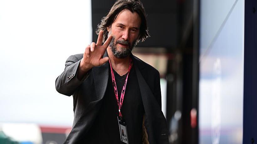 Keanu Reeves waves towards a camera at the 2025 British Grand Prix at Silverstone
