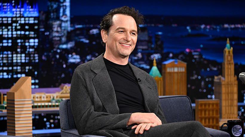 Matthew Rhys, a man with dark-brown hair, wearing a black shirt and greyish blazer is smiling with his hands rested on his leg. The background shows an image of black and orange buildings in the dark. He appears to be a guest on a chat show.