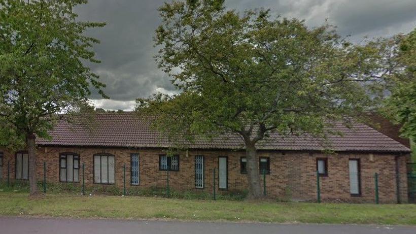 The image shows part of a long single story light-coloured brick building behind a wire fence with two large trees on the verge in front of it.  There are four single-width full length windows and three large double width arched windows, as well as four small windows. The curtains are shut at all the arched windows and two long windows, the remaining two long windows have ornate bars at them.