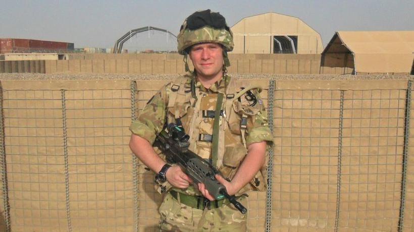 Pete White is standing against an outside wall wearing a soldier's khaki uniform and a helmet and holding a gun across his body.