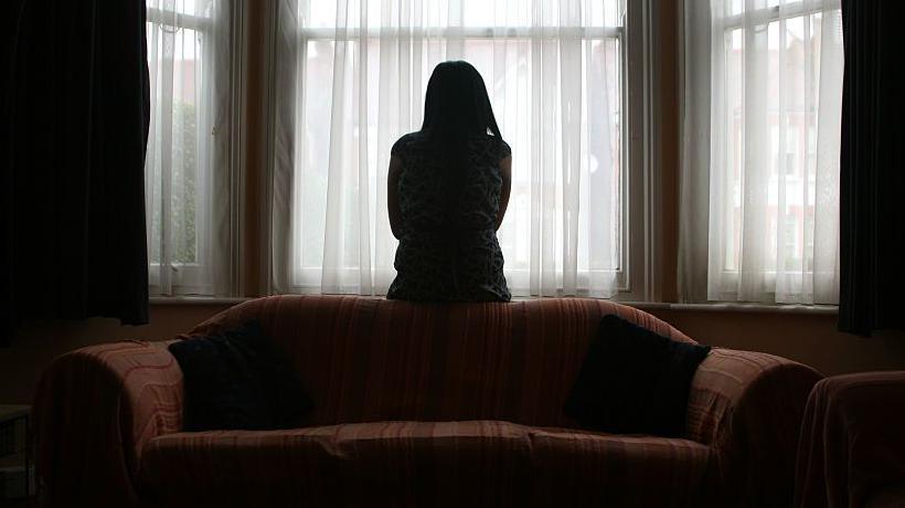 A young woman suffering from domestic violence stands alone in the bay window of her home. 