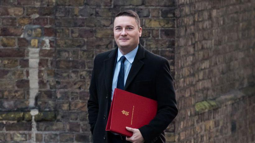 Wes Streeting, a man with short dark hair, wearing a suit and carrying a red folder under his arm.