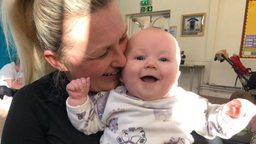 Sian Connel holding her baby sitting down on an exercise mat. Her baby is smiling and holding his hands up in the air. The woman is wearing black and has her blonde hair up in a ponytail. 