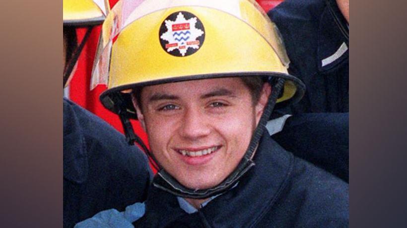 John Alford wearing a fire officer's uniform with a blue jacket and helmet. He is smiling at the camera. 